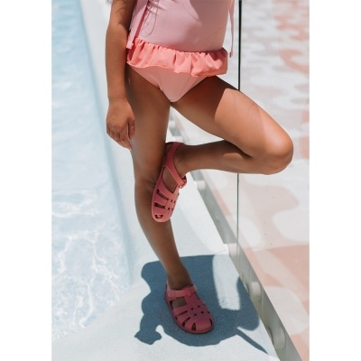 Criança com fato de banho rosa e sandálias rosa ao lado de piscina.