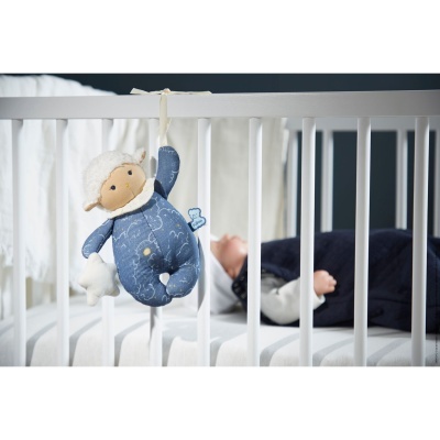 Brinquedo de peluche cordeiro pendurado nas grades de um berço branco com criança deitada ao fundo