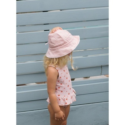 Criança de lado com swimsuit branco de cerejas e chapéu rosa claro, frente a parede azul