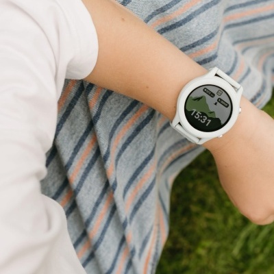 Relógio smartwatch branco com ecrã digital a mostrar 15:31, gráficos e ícones, braço com camisola branca e manta às riscas no fundo.