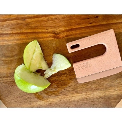 Peça rosa com a marca MAACOM e maçã verde partida sobre tábua de madeira.