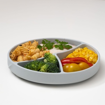 Prato redondo compartimentado com fusilli, vegetais variados numa superfície branca