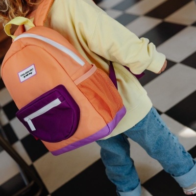 Mochila escolar laranja e roxa com bolso frontal roxo, usada por criança com casaco amarelo e calças azuis em piso xadrez.