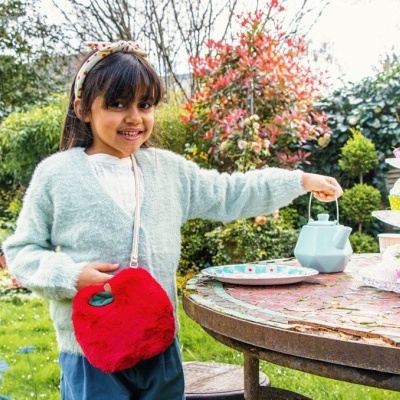 Menina no jardim com regador azul e bolsa vermelha peluda junto de mesa com bolo.