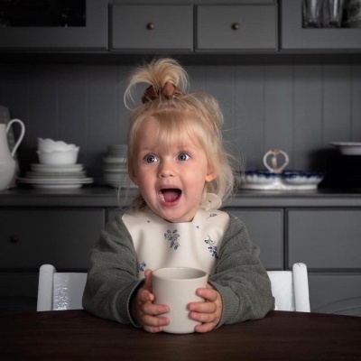 Criança loira segura caneca branca sentada a uma mesa de madeira com armários cinzentos ao fundo