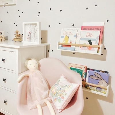 Cadeira rosa com boneca de pano, almofada e cómoda branca junto a parede decorada com pontos pretos e estantes com livros infantis.