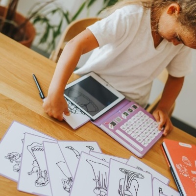 Criança usa tablet com teclado lilás e cartas de desenhos a preto e branco numa mesa de madeira.