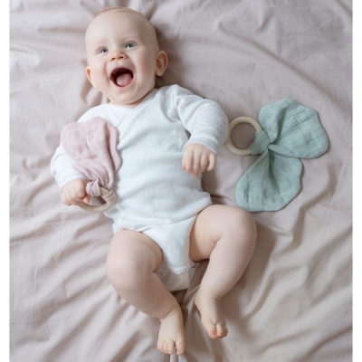 Bebé de body branco deitado segurando brinquedo de tecido com anel de madeira numa cama rosa claro