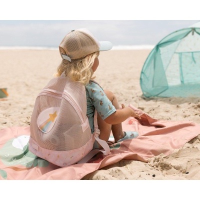 Criança com mochila rosa em forma de canguru sentada na praia sobre toalha cor-de-rosa