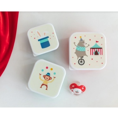 Três caixas brancas com desenhos coloridos de circo e uma chupeta vermelha sobre fundo branco e tecido vermelho