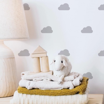 Mantas e toalhas dobradas com brinquedo de peluche de ovelha e blocos de madeira numa mesa ao lado de lâmpada, parede com decalques de nuvens.