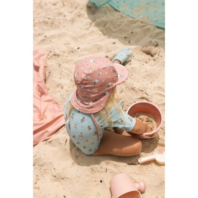 Criança com fato de banho azul e chapéu cor-de-rosa brinca com baldes e pás na areia da praia