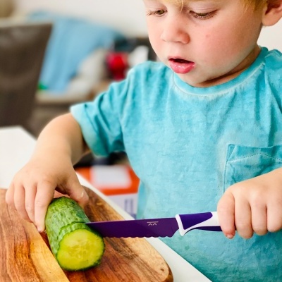 Criança a cortar pepino com faca de lâmina serrilhada roxa na tábua de madeira