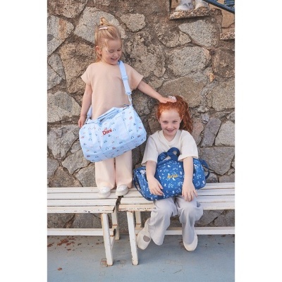 Duas crianças com sacos de maternidade azuis estampados sentadas e em pé em bancos brancos, parede de pedra ao fundo