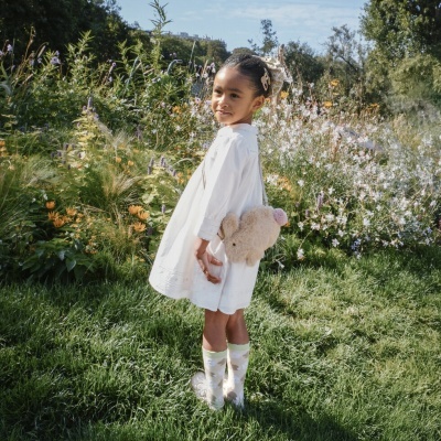Menina com vestido branco, mala de peluche e botas transparentes em campo florido