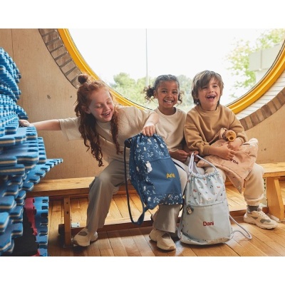 Três crianças sorridentes sentadas num banco de madeira junto a uma janela redonda, segurando mochilas personalizadas.