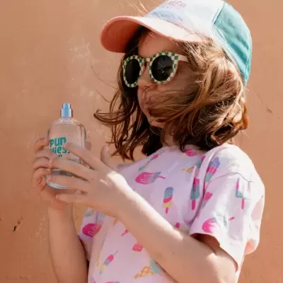 Menina com óculos quadrados, boné colorido e t-shirt de gelados segurando frasco spray transparente com texto azul