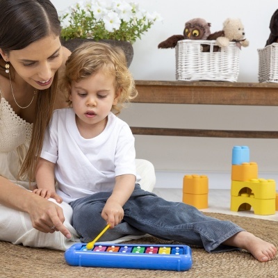 Criança a tocar xilofone azul com mulher ao lado e blocos coloridos no chão