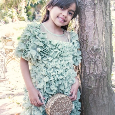 Menina em vestido verde com mala redonda dourada junto a árvore