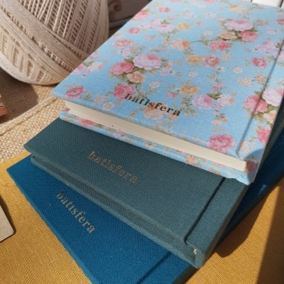 Três cadernos com capas de tecido em diferentes tons de azul e a palavra 'baustera' impressa, sobre superfície amarela junto a novelo de linha.