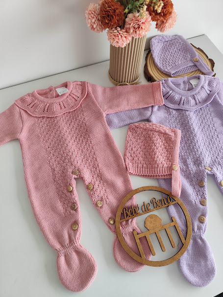 Macacões de bebé rosa e lilás de malha com botões e gorros, decorados com flores, placa de madeira na mesa