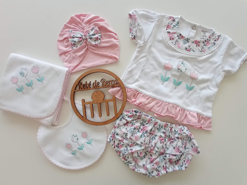 Conjunto de roupa para bebé com flores, touca rosa, babete e toalha bordados, sobre superfície branca