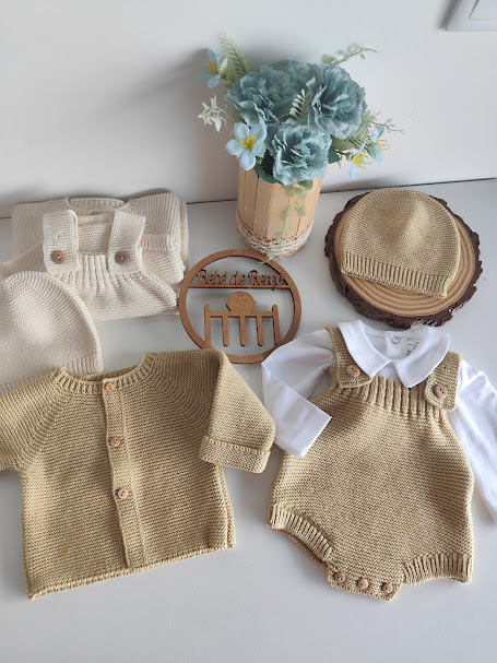 Conjunto de roupa de bebé em malha bege claro com cardigã, jardineira, gorro e calças junto a vaso de flores artificiais