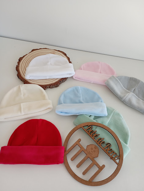 Seis gorros de bebé de várias cores sobre superfície branca com suporte redondo de madeira e círculo de madeira decorativo