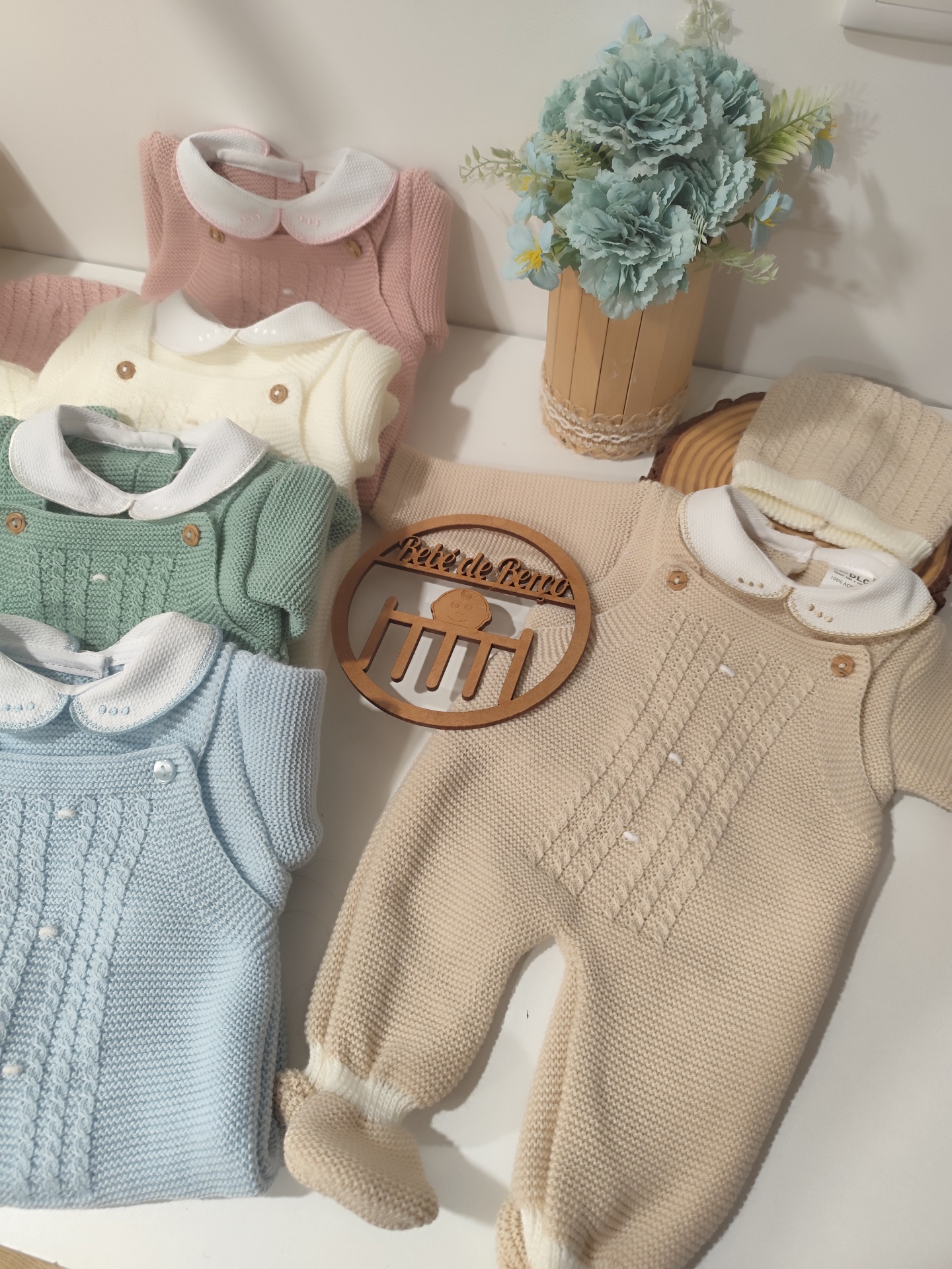 Conjunto de roupa de bebé em malha de várias cores dispostas com vaso de flores ao lado.