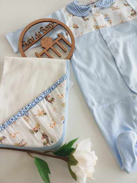 Conjunto de roupa de bebé azul e cobertor branco com detalhes em padrão vichy e desenhos animados, com ornamento de madeira e flor branca