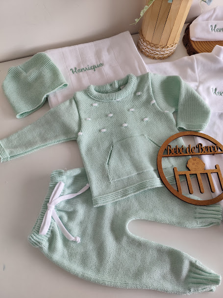 Conjunto de roupa de bebé verde claro em malha com camisola, calças e gorro, sobre fraldas brancas bordadas com o nome Henrique