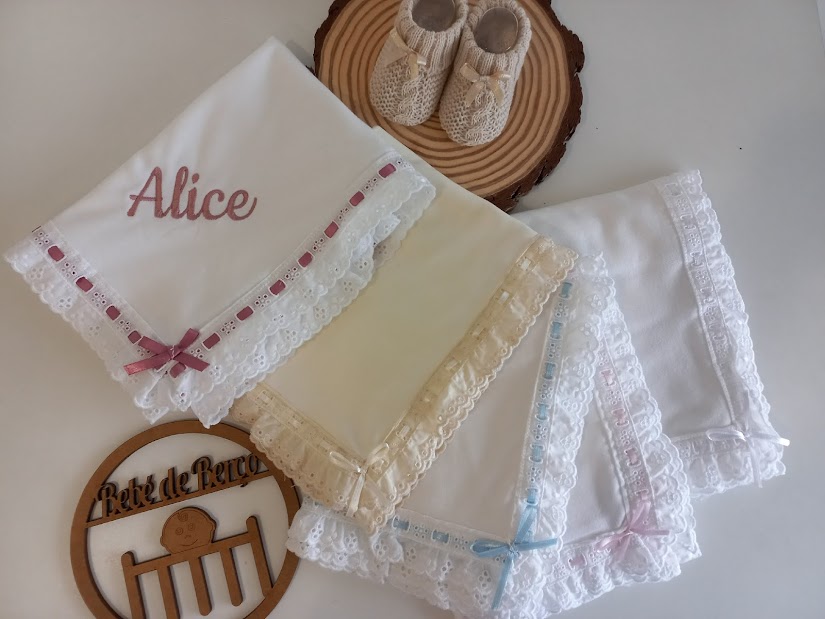Quatro toalhas ou fraldas de pano com bordados em renda e laços coloridos, umas sapatilhas bege e suporte de madeira.