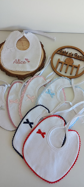 Babetes brancos bordados com laços coloridos e um com o nome Alice, ao lado de enfeite de madeira