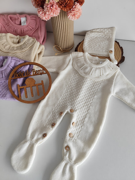 Macacão de bebé branco em malha com botões e gola tricotada, gorro e outros macacões coloridos ao fundo
