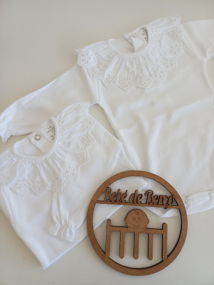 Dois bodies brancos de bebé com golas rendadas e placa em madeira com texto 'Bebé de Berço'