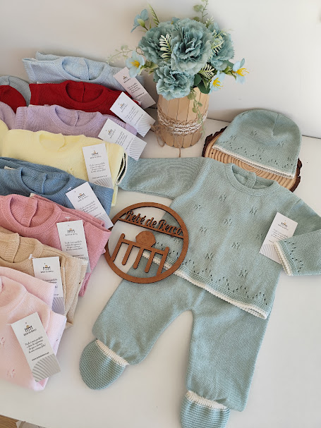 Conjunto de roupa de bebé azul esverdeado com padrão e gorro em mesa com flores e mais conjuntos coloridos