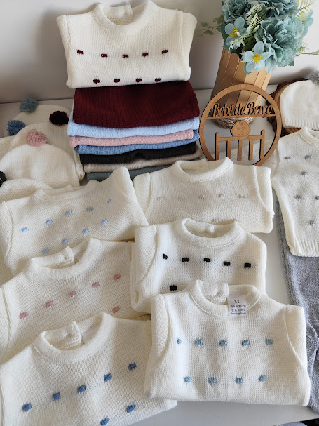 Suéteres infantis de malha em branco com bordados coloridos e um chapéu com pompons ao lado de uma placa de madeira com o nome BebedeBebé