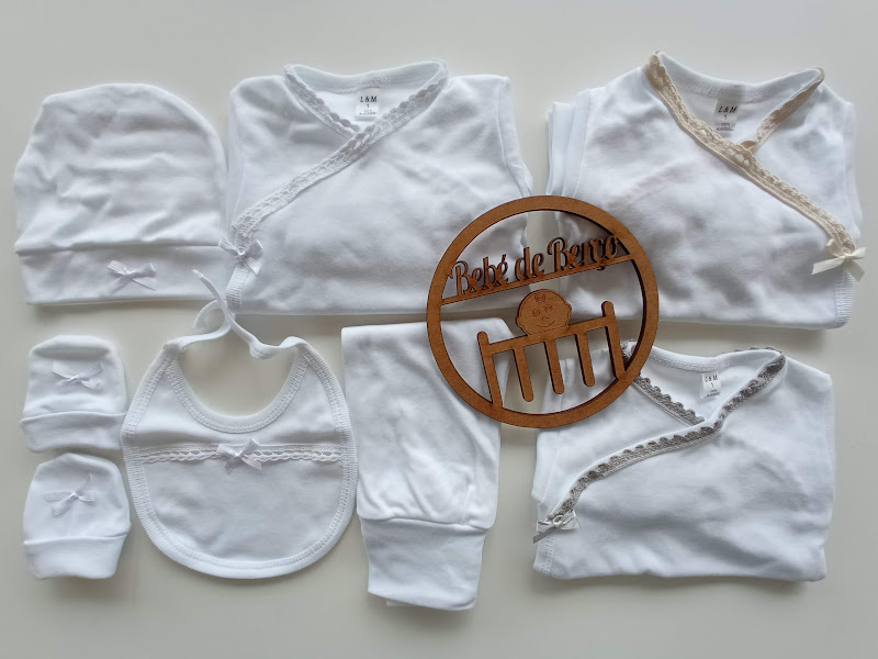 Conjunto de roupa de bebé branca com detalhes em fita e renda, e placa redonda de madeira