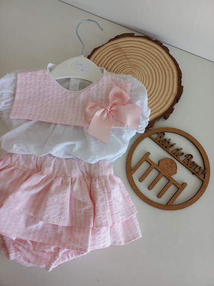 Conjunto de roupa de bebé rosa e branco com laço, pendurado em cabide, com ornamentos em madeira ao lado.