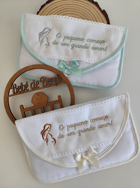 Duas bolsas de pano branco bordadas com laços e mensagem em português, sobre uma superfície clara com ornamento de madeira