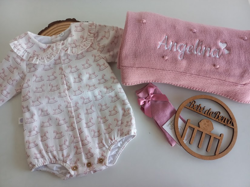 Conjunto de bebé com body branco estampado, manta rosa tricotada, faixa de cabelo rosa e placa em madeira