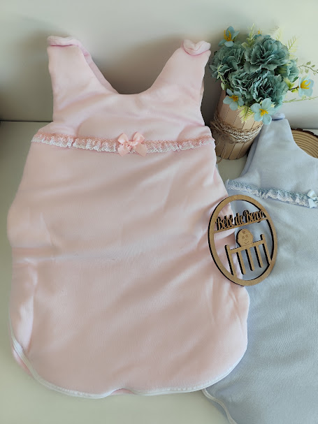 Dois sacos de dormir para bebé em rosa claro e azul claro com detalhes em renda, junto a uma planta decorativa e emblema de madeira BD LUX BABY