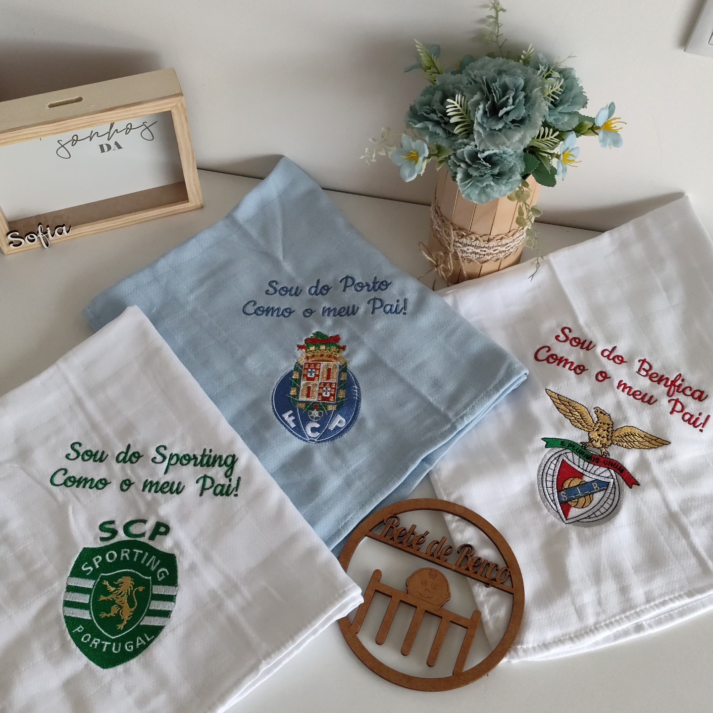 Três toalhas bordadas com emblemas dos clubes Sporting, Porto e Benfica junto a vaso de flores e objetos decorativos de madeira