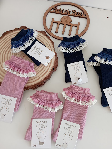 Meias de bebé rosa e azul com folhos rendados e etiquetas com texto sobre superfície branca e base de madeira