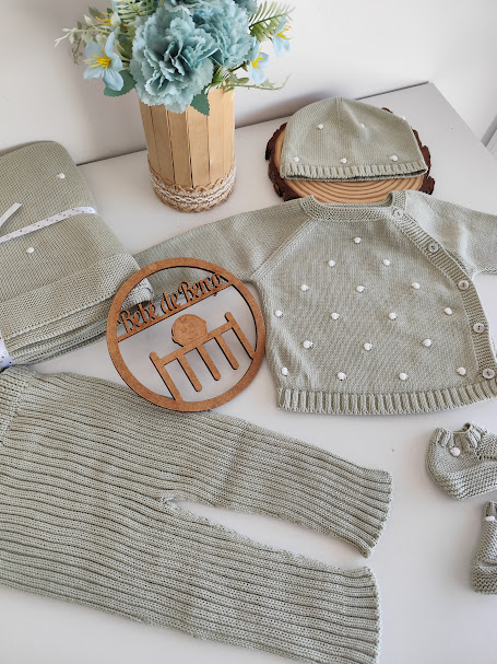 Conjunto de roupa de bebé em malha bege com detalhes brancos e acessórios sobre fundo branco.