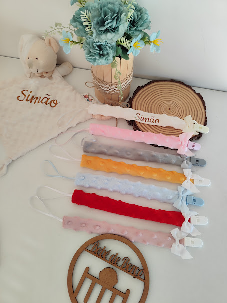 Fitas porta-chupetas coloridas e texturizadas com molas e laços, decoradas num fundo branco com peluche e flores artificiais.