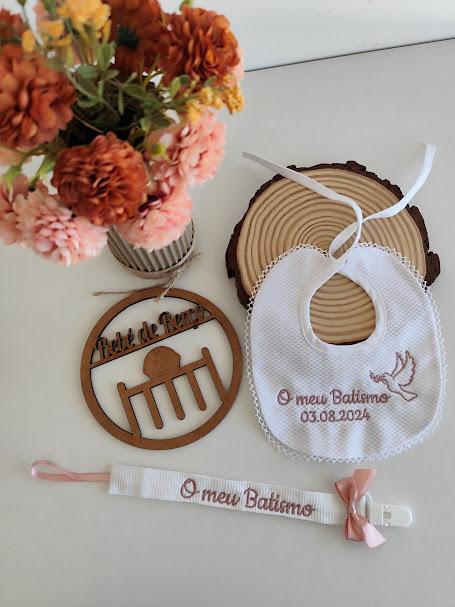 Babete branco, prendido de chupeta e placa de madeira com tema de batismo ao lado de vaso com flores