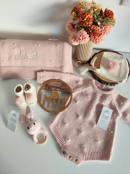 Conjunto de bebé rosa claro com body, touca, sapatos, manta bordada com nome, brinquedo de madeira e flores decorativas.