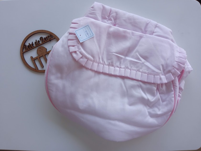 Colcha rosa claro para bebé com pregas decorativas sobre superfície branca e círculo decorativo de madeira ao lado