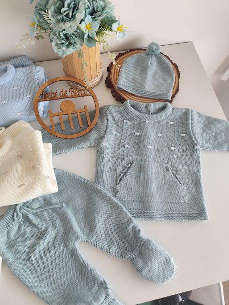 Conjunto de roupa de bebé de malha azul claro com casaco, calças e gorro, placa de madeira e vaso com flores